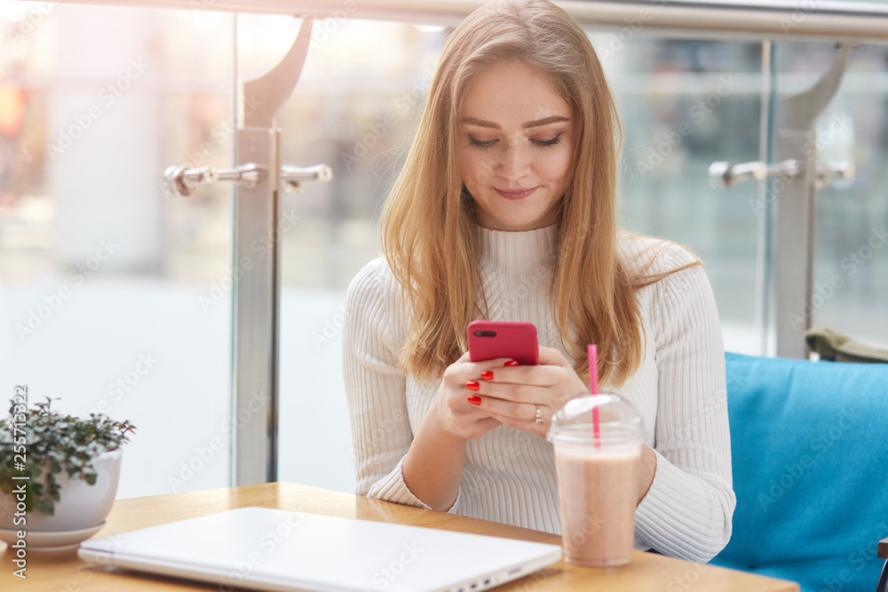Elegant young blonde woman in white sweater sits on blue sofa in cafe, holds pink mobile phone and surfes internet or types mesage to her friend. Attractive woman drinks cocktail and check email. Elegant young blonde woman in white sweater sits on blue sofa in cafe, holds pink mobile phone and surfes internet or types mesage to her friend. Attractive woman drinks cocktail and check email.