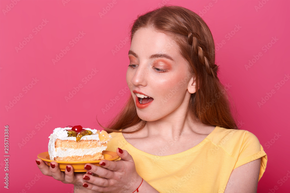 Attractive young woman with opened mouth holds plate with piece of delicious cake in hands. Brown haired lady with red manicure poses in studio isolated over pink background. Sweet food concept. Attractive young woman with opened mouth holds plate with piece of delicious cake in hands. Brown haired lady with red manicure poses in studio isolated over pink background. Sweet food concept.