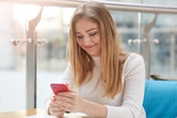 Smiling young blonde woman with long straight hair, in white shirt massages in her cellphone while sitting in cafe. Attractive girl recives sms from her boyfriend, has pleasent news. People concept.