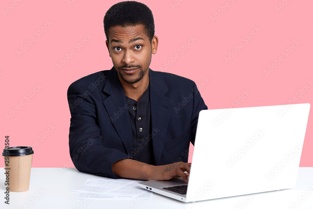 Indoor portrait of young handsome black man sitting at table, drinks coffee or tea and works with lap top. Model looks surprised at camera while typing on keyboard, expreses astonishment with face. Indoor portrait of young handsome black man sitting at table, drinks coffee or tea and works with lap top. Model looks surprised at camera while typing on keyboard, expreses astonishment with face.