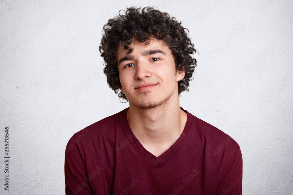 Close up shot of curly haired European teenager with little beard, wears maroon shirt. Young smiling male stands with lowered shoulders against white studio wall, looks shy. Model poses in studio. Close up shot of curly haired European teenager with little beard, wears maroon shirt. Young smiling male stands with lowered shoulders against white studio wall, looks shy. Model poses in studio.