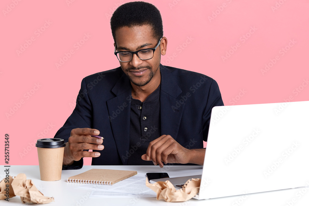 Portrait of dark skinned man in black jacket and spectacles works with laptop, writes notes in notebook and drinks coffee. Handsome black male sits at desk, looks thoughtful. Online working concept. Portrait of dark skinned man in black jacket and spectacles works with laptop, writes notes in notebook and drinks coffee. Handsome black male sits at desk, looks thoughtful. Online working concept.