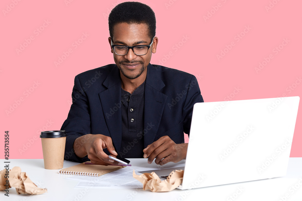 Handsome African American male sits at desk with lap top and crumpled shirts of paper, works online. Dark skinned guy takes smart phone and looks down on it, drinks coffee or tea. Technology concept. Handsome African American male sits at desk with lap top and crumpled shirts of paper, works online. Dark skinned guy takes smart phone and looks down on it, drinks coffee or tea. Technology concept.