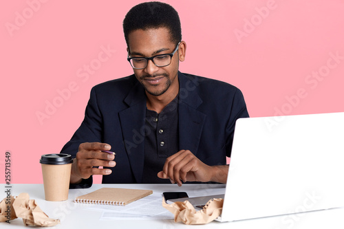 Portrait of dark skinned man in black jacket and spectacles works with laptop, writes notes in notebook and drinks coffee. Handsome black male sits at desk, looks thoughtful. Online working concept. photo