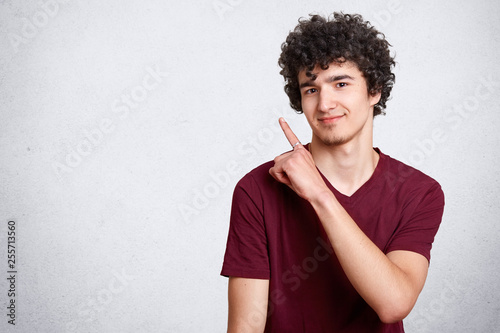 Portrait of stylish handsome young teenager isolated on white background. Dark haired curly student smiles and points aside. Attractive teen poses in photo studio. Copy space for your advertsment. photo