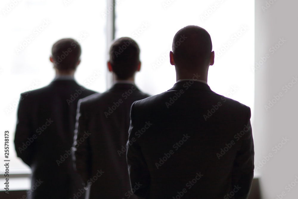 rear view of three businessmen as they stare at the big window overlooking the city rear view of three businessmen as they stare at the big window overlooking the city