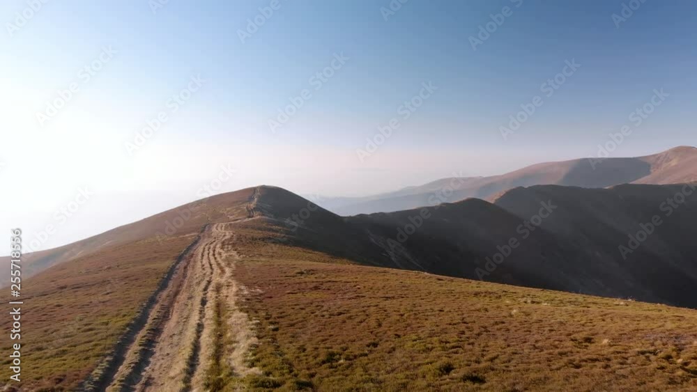 Aerial drone flying backwards while man hiker walking on mountain road with distant valley peak on horizon at sunrise Aerial drone flying backwards while man hiker walking on mountain road with distant valley peak on horizon at sunrise