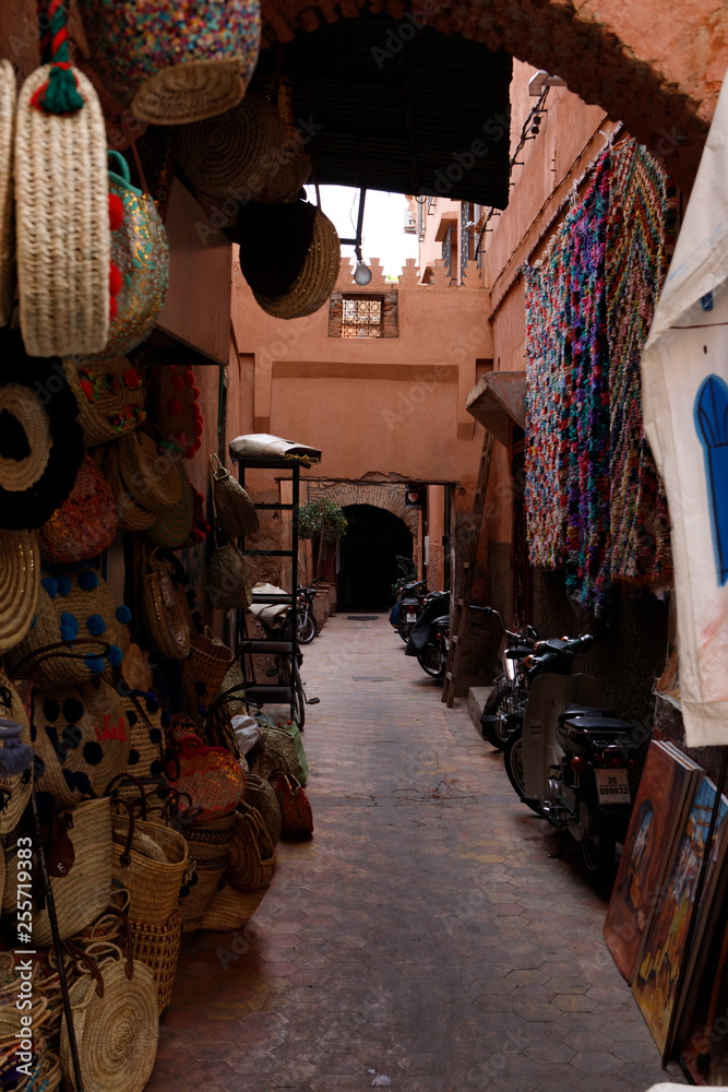 Naklejka premium Typical Moroccan alley in the souks of the Medina of Marrakesh displaying scarfs, dresses and other craft products (Marrakesh, Morocco, Africa)
