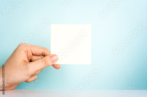 Human hand holding a beige blank note on the office desk and blue background. Design announcement message concept at work.