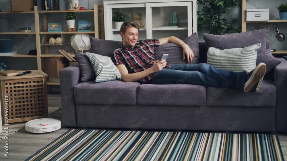 Smiling guy is sufring the Internet with smartphone relaxing on sofa while robotic vacuum cleaner is cleaning floor removing dust from carpet. Gadgets and housekeeping concept.