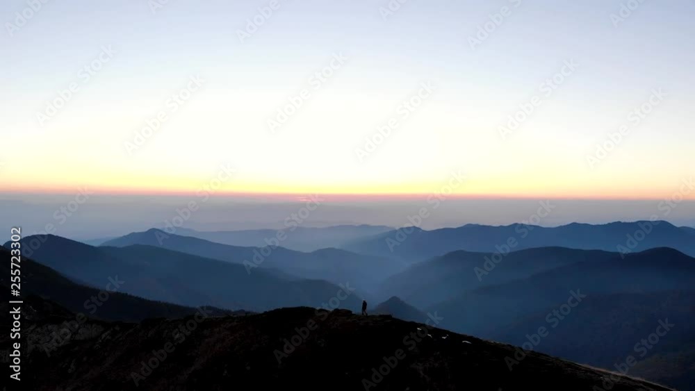 Drone aerial flying forward over mountain ridge with man hiker walking on top, looking into picturesque valley of endless blue silhouettes at sunset Drone aerial flying forward over mountain ridge with man hiker walking on top, looking into picturesque valley of endless blue silhouettes at sunset