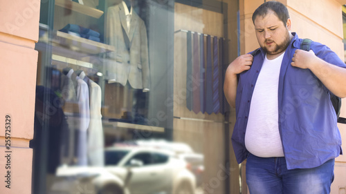 Obese man sadly looking at suits in shop window, overweight problem, motivation
