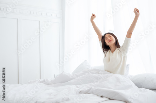 Asian woman wake up in the morning, sitting on white bed and stretching, feeling happy and fresh