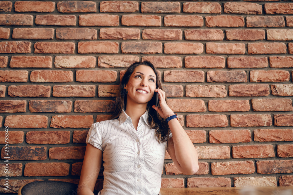 Smiling Caucasian pretty woman leaning on the brick wall in cafeteria and using smart phone. Smiling Caucasian pretty woman leaning on the brick wall in cafeteria and using smart phone.