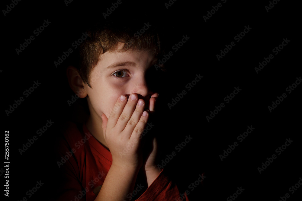 Portrait in the dark of a young scared sad boy of 7 years scared and upset hands on the mouth in the dark backround in the profile. Portrait in the dark of a young scared sad boy of 7 years scared and upset hands on the mouth in the dark backround in the profile.