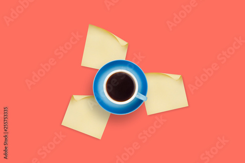 Workplace on table of coral color with one blue ceramic cup full of black coffee on saucer and blank square paper stickers with curved corner. Top view. Office work concept