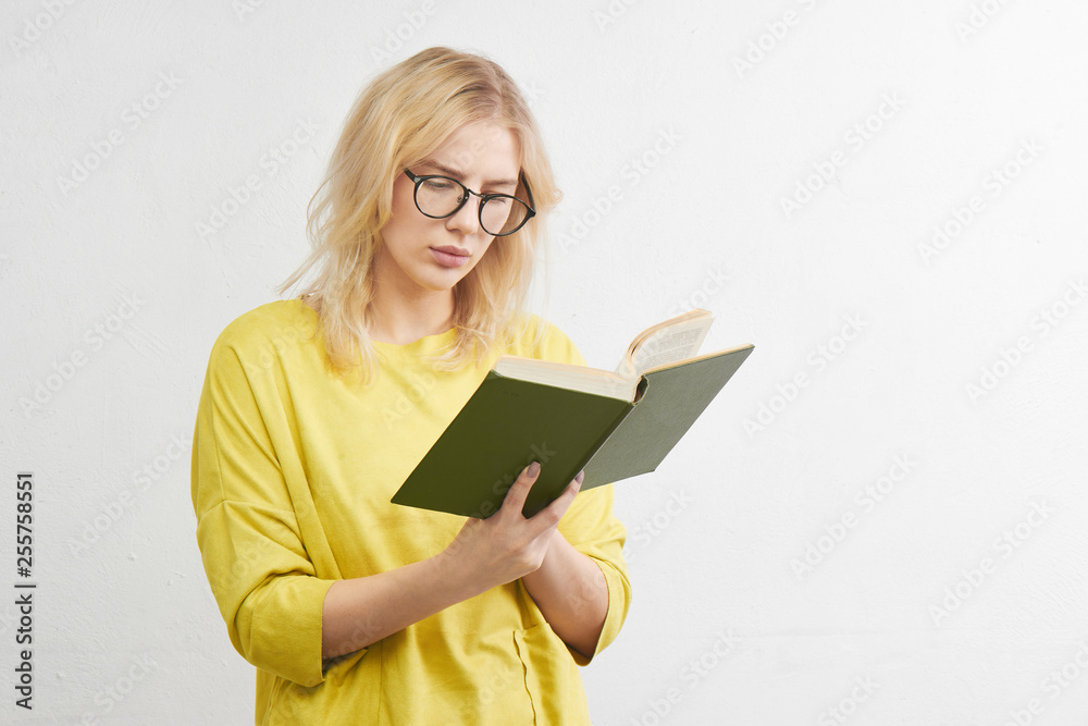Smart young blonde with glasses reads a book with a serious face in yellow clothes alone in a white studio background Smart young blonde with glasses reads a book with a serious face in yellow clothes alone in a white studio background