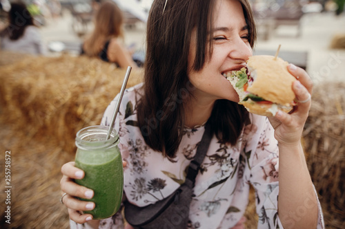 Stylish hipster girl in sunglasses eating delicious vegan burger and holding smoothie in glass jar in hands at street food festival. Happy boho woman biting burger with drink in summer street photo