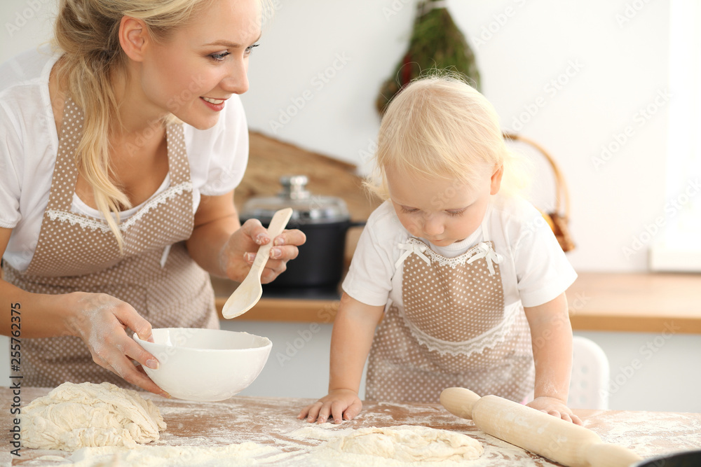 Little girl and her blonde mom in beige aprons playing and laughing while kneading the dough in kitchen. Homemade pastry for bread, pizza or bake cookies. Family fun and cooking concept Little girl and her blonde mom in beige aprons playing and laughing while kneading the dough in kitchen. Homemade pastry for bread, pizza or bake cookies. Family fun and cooking concept