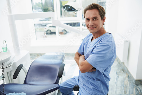 Joyful male stomatologist posing near dental chair