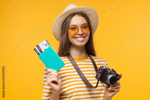 Excited cheerful young tourist girl holding passport with tickets and camera, isolated on yellow background