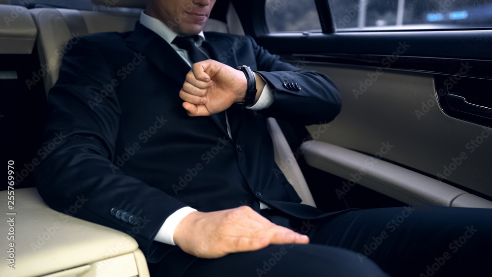 Businessman riding on backseat in luxury car, looking at watch, late for meeting Businessman riding on backseat in luxury car, looking at watch, late for meeting