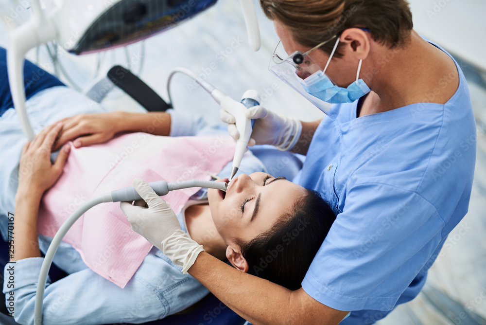 Competent dentist in protective mask doing teeth cleaning for young lady Competent dentist in protective mask doing teeth cleaning for young lady