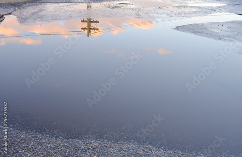 Puddle water reflection of sky clouds and power line. Above ground electricity pole reflected in urban setting background. Parking lot with abstract water puddle reflection
