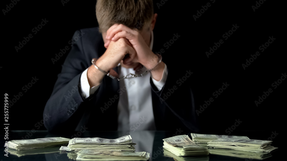 Upset businessman in handcuffs, money lying on table, corruption frauds, bribe Upset businessman in handcuffs, money lying on table, corruption frauds, bribe
