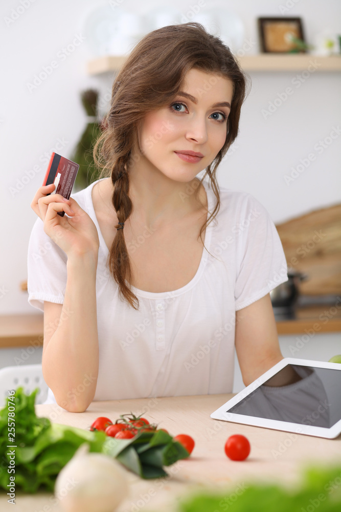 Young woman looking for a new recipe for cooking in a kitchen. Housewife is making online shopping by tablet computer and credit card Young woman looking for a new recipe for cooking in a kitchen. Housewife is making online shopping by tablet computer and credit card