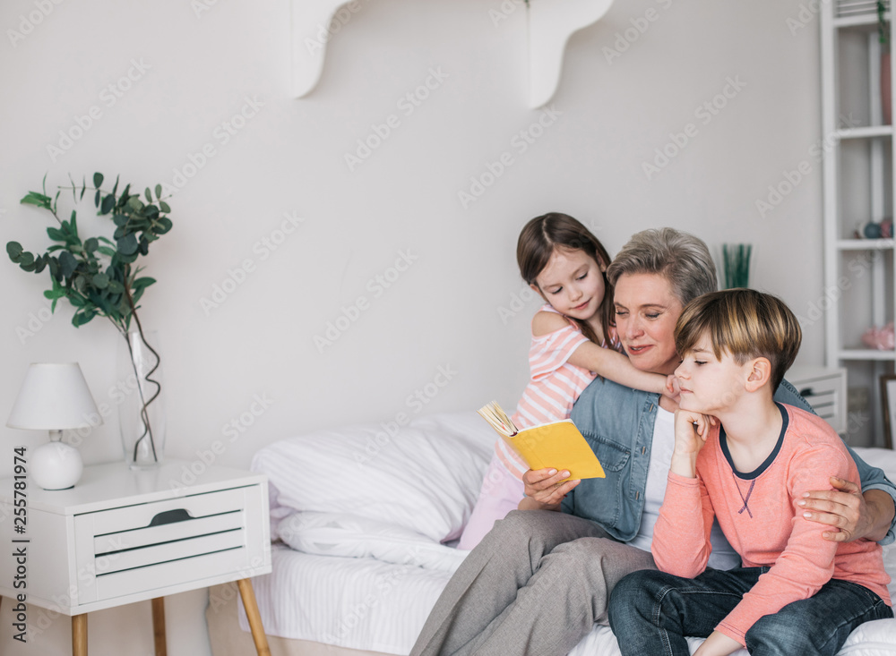 Cropped photo of gray-haired woman reading book for her grandchildren in house