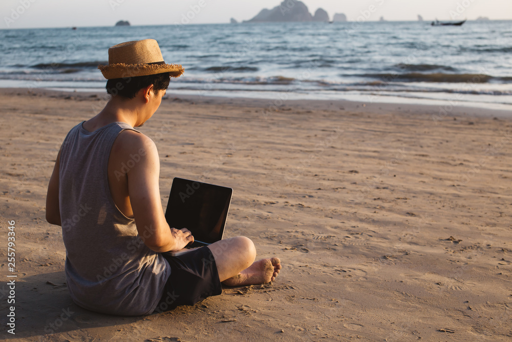 Back view of barefoot man in hat sitting on sandy beach near waving sea and browsing modern laptop