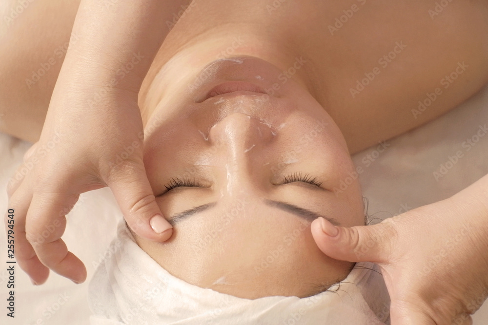 Naklejka premium Portrait of Asian woman relaxes on a sofa with eyes closed in a towel on her head. The hands of a cosmetologist are massaging the eyebrows at the face of a woman in a beauty salon. Cosmetology.