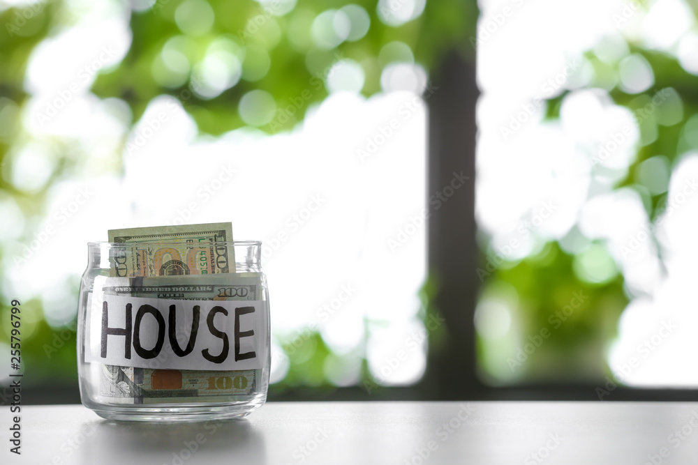 Glass jar with money and word HOUSE on table against blurred background, space for text