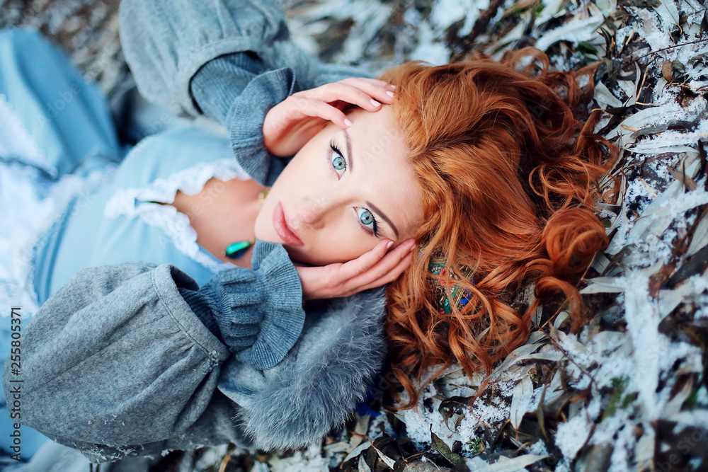 Foto de Beautiful young girl of European appearance with red hair and blue  eyes lying on the snow in spring Park. Bright charming woman with a deep  look in an old dress.