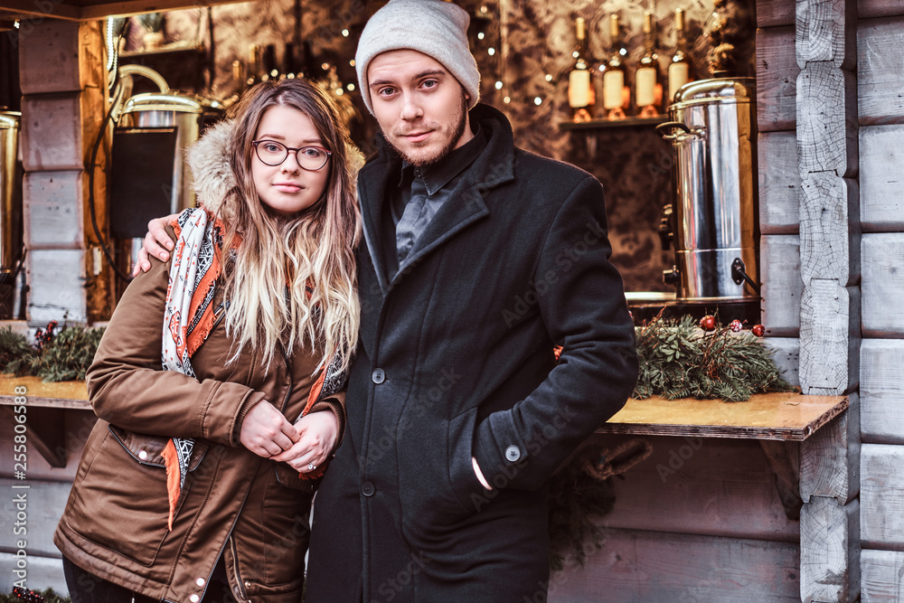 Portrait of a young couple, hugging and looking at the camera while standing near the counter with souvenirs at the winter Christmas Fair