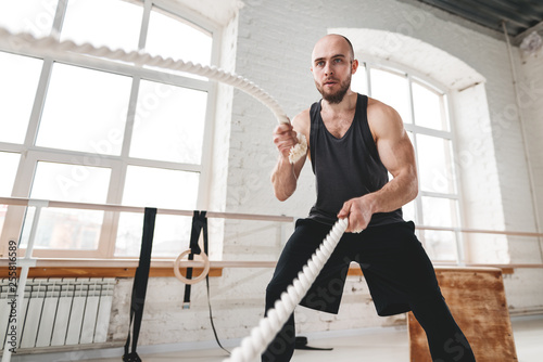 Strong man workout with battle ropes at light gym. Muscular sportsman doing crossfit excursion with ropes in workout gym photo