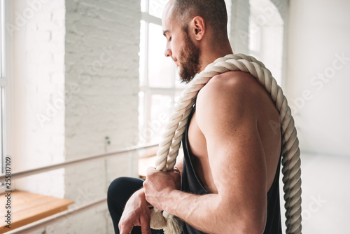 Handsome muscular man holding on shoulder rope workout in light gym. Portrait of fitness man with battle ropes at cross gym