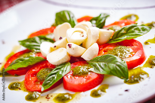 Traditional Italian caprese salad with sliced tomatoes, mozzarella cheese, basil and pesto sauce on white plate. Close up