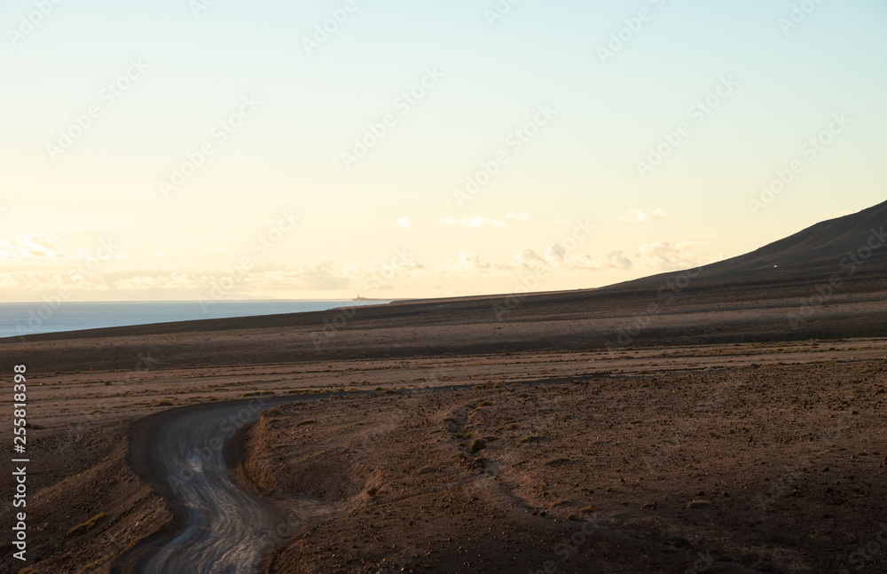 Dirt road to Cofete beach at sunset. Fuerteventura, Canary Islands, Jandia natural park. Ocean and high hills in the background. Dirt road to Cofete beach at sunset. Fuerteventura, Canary Islands, Jandia natural park. Ocean and high hills in the background.