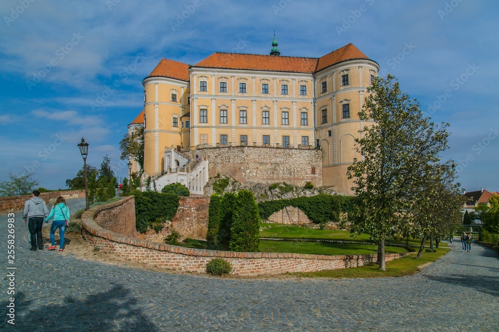 Mikulov, Czech Republic / South Moravia - October 15 2016: Mikulov castle with yellow and white facade and red roof standing on a rock, green vegetation in garden, tourists walking on pavement