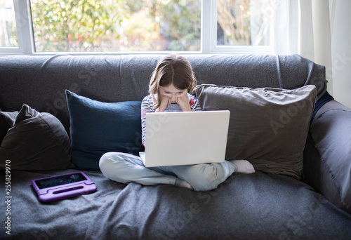 female child sitting on the sofa at home with a laptop and head phones