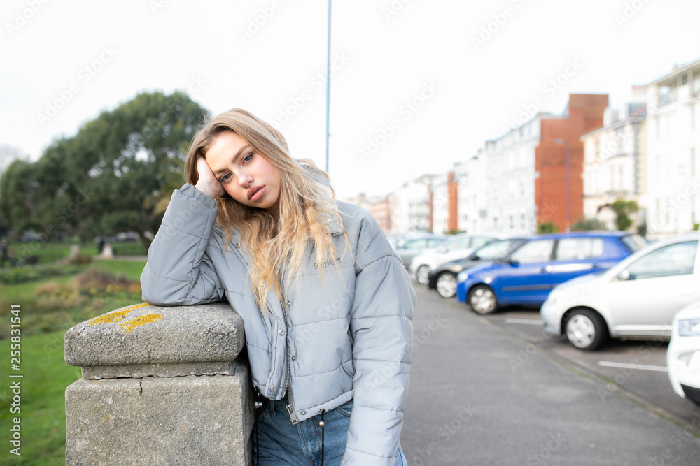 Teenage girl leaning on a concrete post on a walk way with cars parked