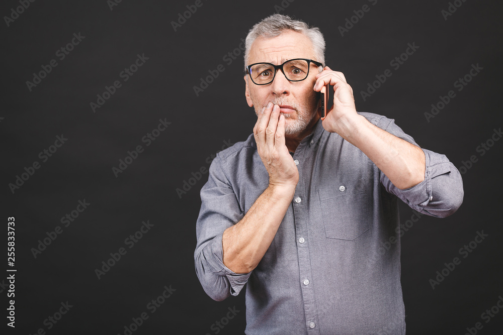 Waist-up shot of shocked senior man with glasses, worry and surprise holding smartphone receiving bad news looking concerned and stunned at camera isolated over black background. Waist-up shot of shocked senior man with glasses, worry and surprise holding smartphone receiving bad news looking concerned and stunned at camera isolated over black background.