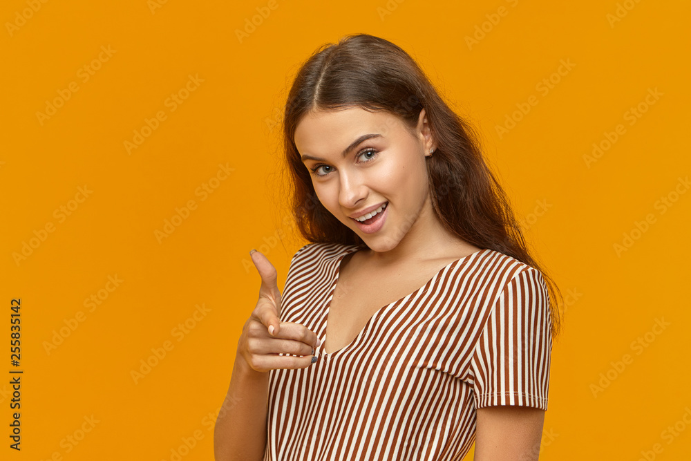 Cunning gorgeous young Caucasian woman dressed in striped top looking at camera with excited smile, pointing fore finger at you. Human facial expressions, gestures, signs and body language