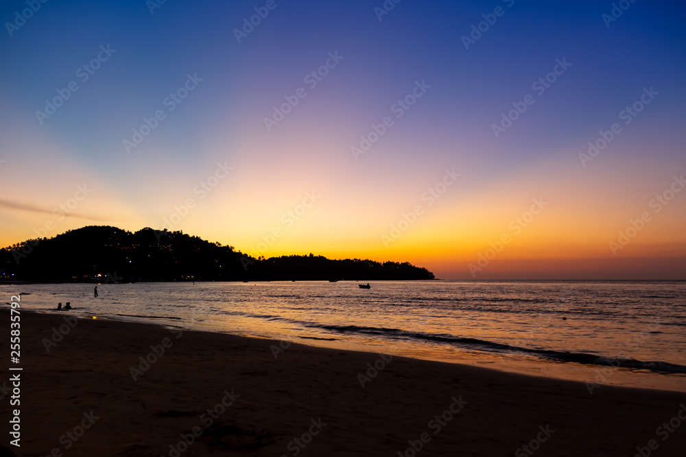 People enjoy the sunset on the sandy beach. Beautiful setting sun in blue-orange tones against the background of the mountain. People enjoy the sunset on the sandy beach. Beautiful setting sun in blue-orange tones against the background of the mountain.