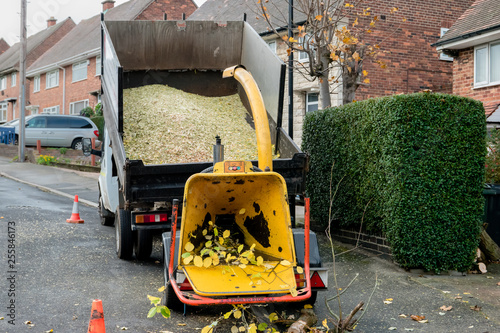 An engine-powered drum woodchipper towed behind a tipper dump truck, showing the hopper and discharge chute with wood chips photo