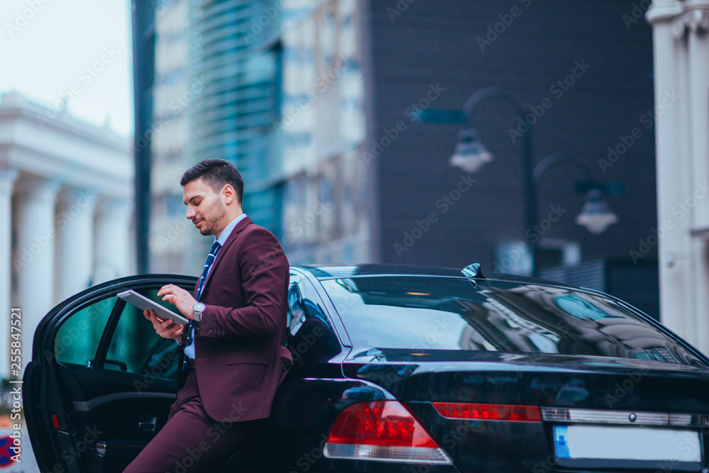 Handsome young businessman leaning on his limo and smiling while looking at his phone. Handsome young businessman leaning on his limo and smiling while looking at his phone.