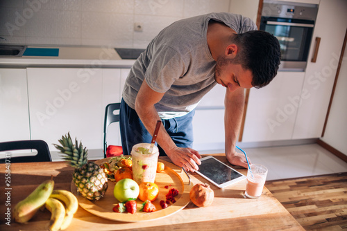 Vegan man in his 20's has a morning preparation of a smoothie while looking at a recipe on his tablet.