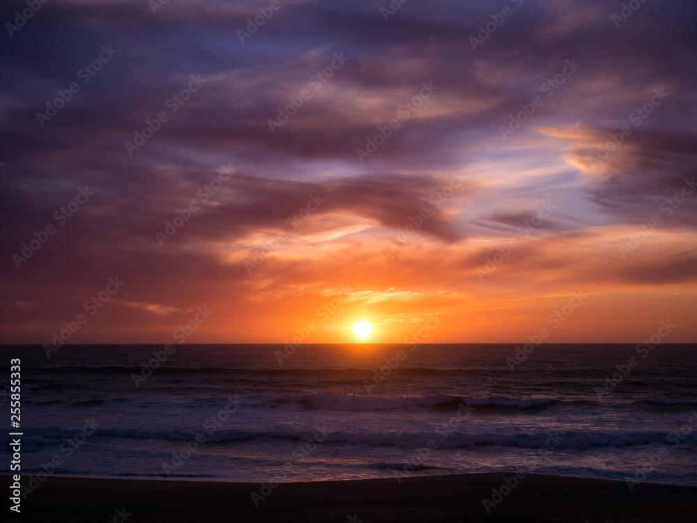 Sun on horizon at sunset with blue orange gradient sky and dark dramatic clouds over ocean